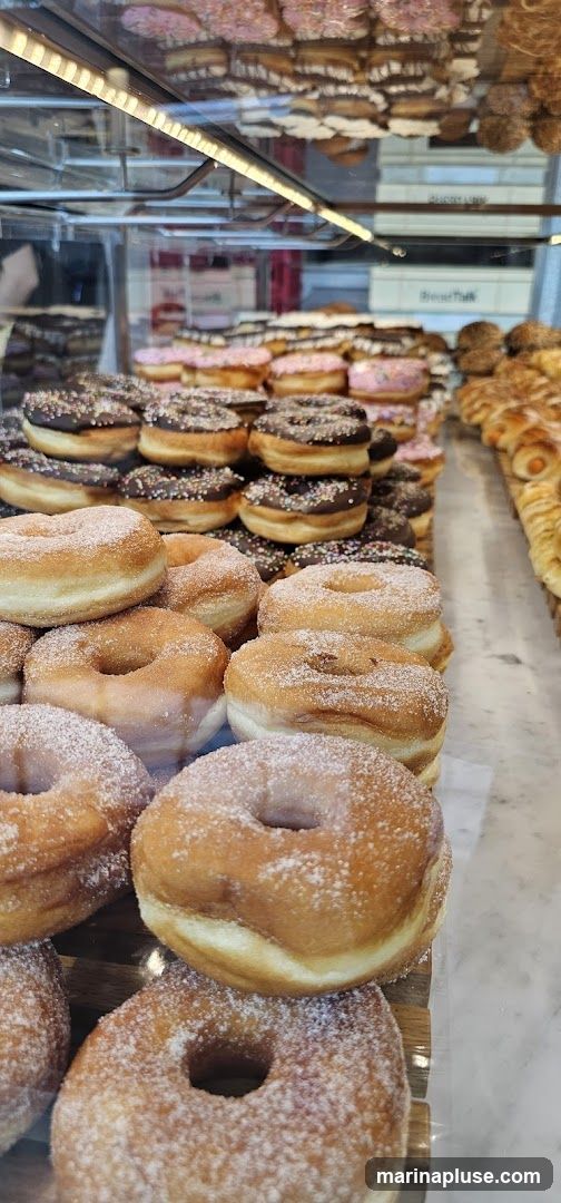 A close-up view of a bakery display case filled with various donuts and pastries. In the foreground, several plain donuts coated in sugar are stacked. Behind them, donuts with chocolate frosting and sprinkles are visible, along with pink frosted donuts. To the right, a row of savory pastries, possibly filled croissants or sausage rolls, can be seen. The display is illuminated by overhead lighting, highlighting the textures and colors of the baked goods. In the background, through the glass, a building facade with signage is partially visible.