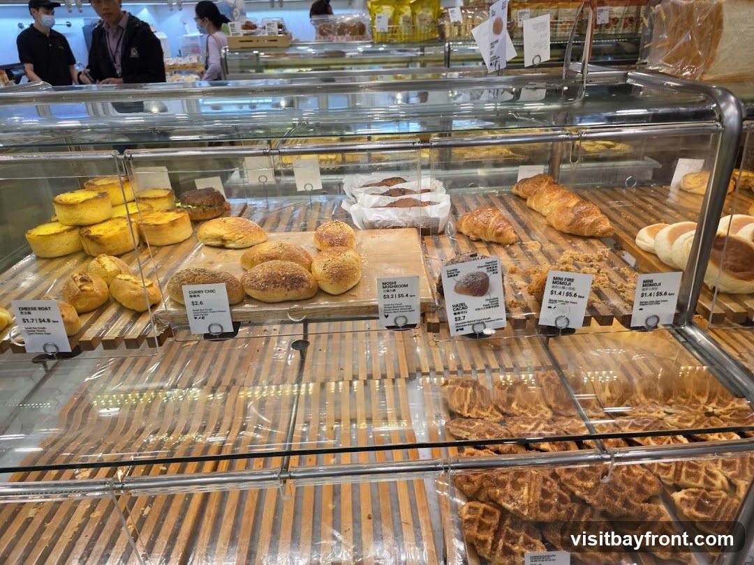 A display case filled with various baked goods, including tarts, buns, croissants, and waffles. Several labels with product names and prices are visible. In the background, customers and staff can be seen.