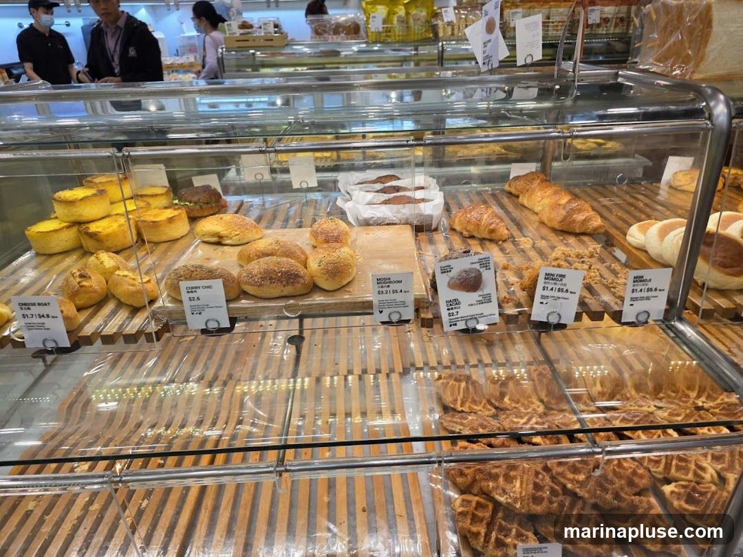 A display case filled with various baked goods, including tarts, buns, croissants, and waffles. Several labels with product names and prices are visible. In the background, customers and staff can be seen.