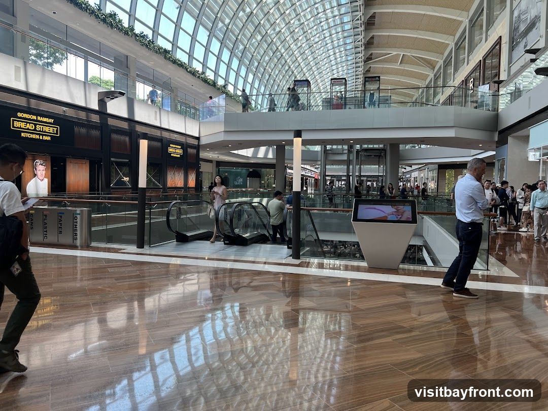 The image shows the interior of a large, modern shopping mall with a distinctive glass-domed ceiling. In the foreground, people are walking on a polished wooden floor. In the mid-ground, there are escalators and a digital display screen. On the upper level, a restaurant with signage for "GORDON RAMSAY BREAD STREET KITCHEN & BAR" is visible. Other shops and shoppers are present on various levels.
