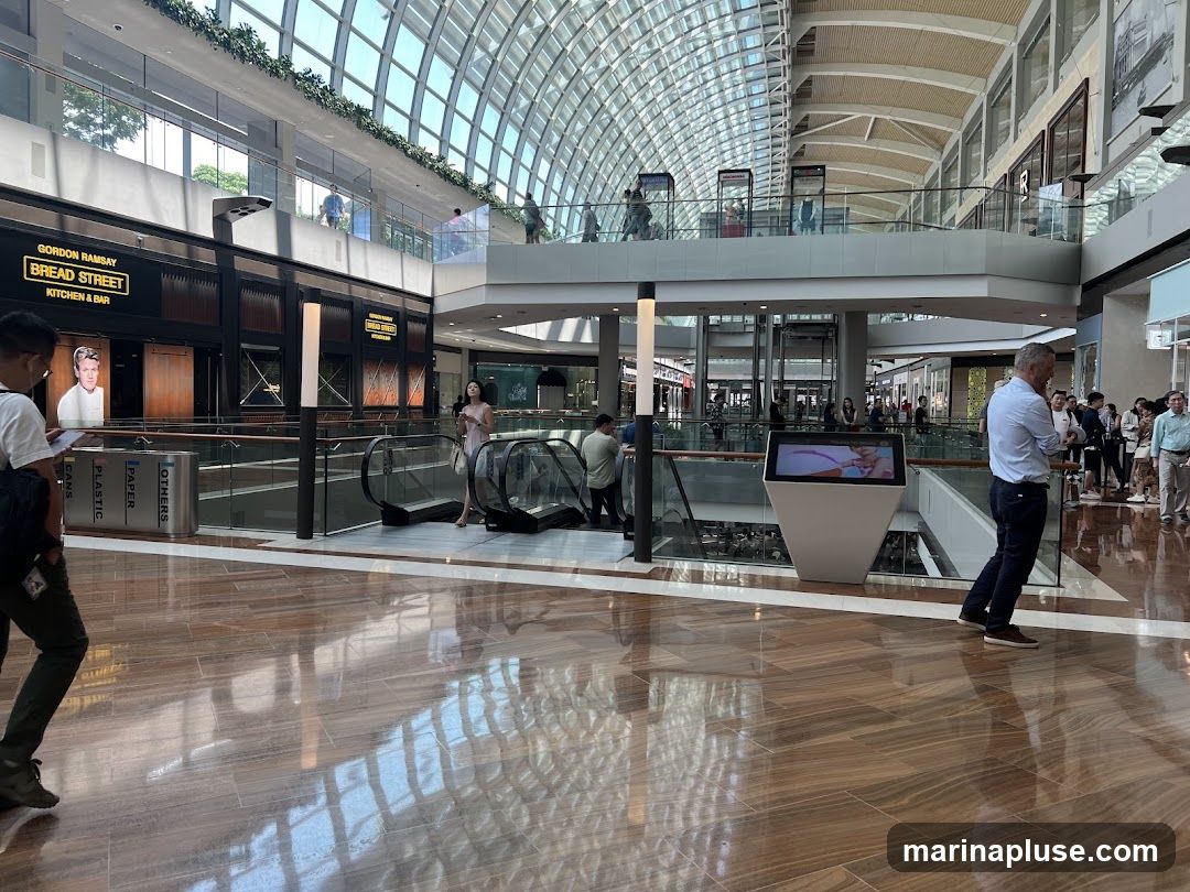 The image shows the interior of a large, modern shopping mall with a distinctive glass-domed ceiling. In the foreground, people are walking on a polished wooden floor. In the mid-ground, there are escalators and a digital display screen. On the upper level, a restaurant with signage for "GORDON RAMSAY BREAD STREET KITCHEN & BAR" is visible. Other shops and shoppers are present on various levels.
