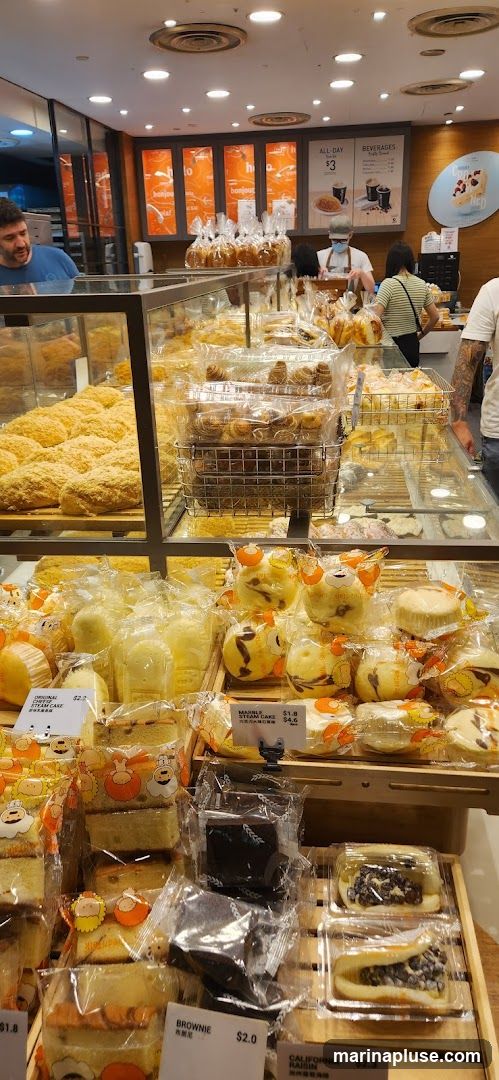 A variety of baked goods are displayed in a bakery. In the foreground, there are individually packaged cakes and brownies. Further back, larger loaves of bread and pastries are visible in display cases. A menu board with beverage options and an all-day set is visible in the background.