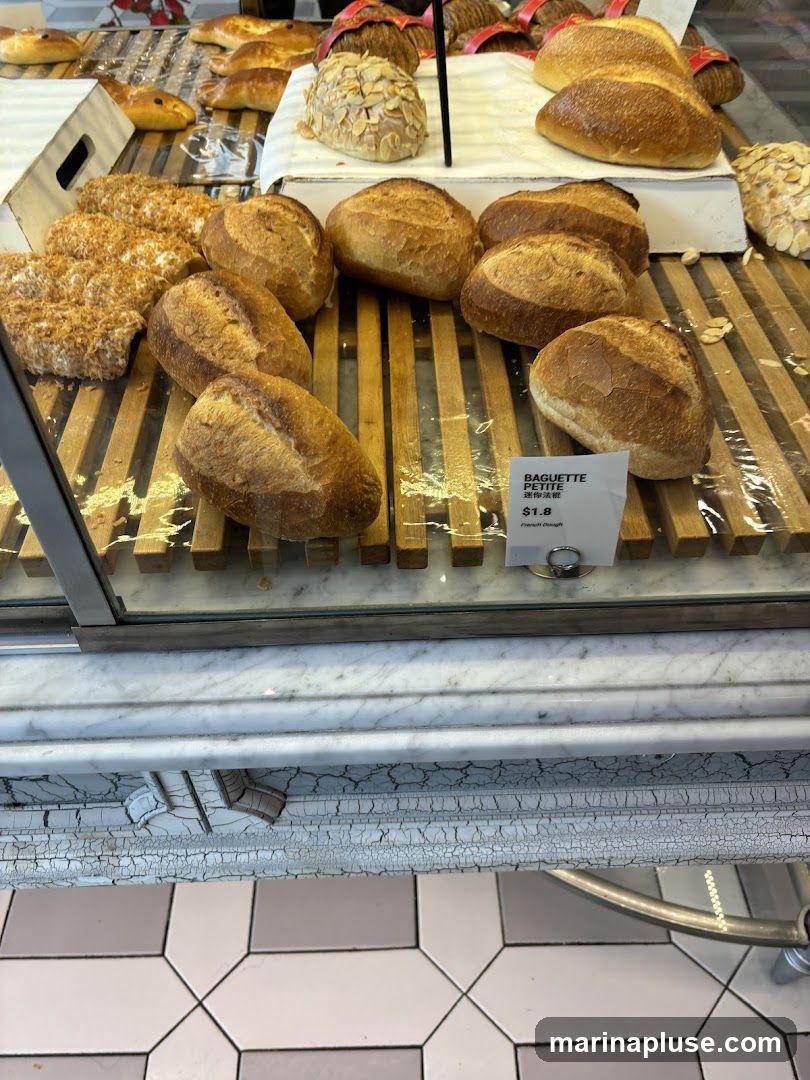 A display of various baked goods, including loaves of bread, baguettes, and pastries, arranged on wooden racks behind a glass counter. A small sign indicates 'BAGUETTE PETITE' for $1.8, described as 'French Dough'.