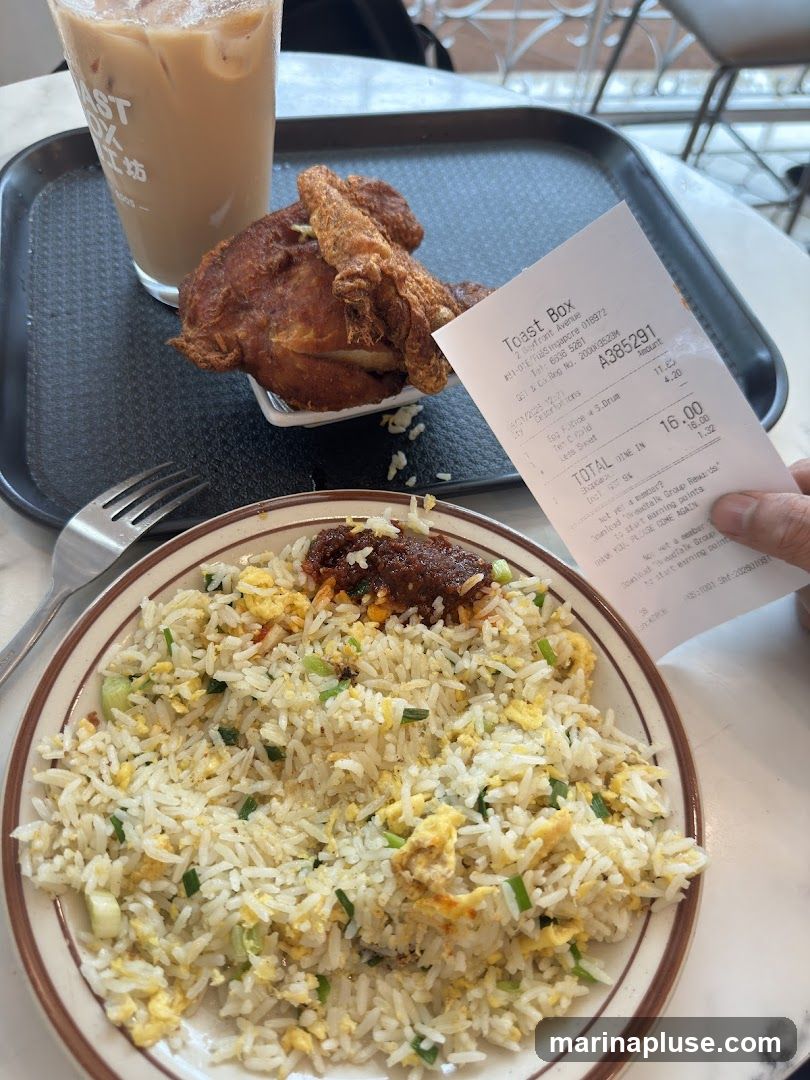 A close-up view of a meal consisting of fried rice, fried chicken, and a beverage, placed on a black tray. A receipt from "Toast Box" is held in a person's hand, showing the total cost of the meal.