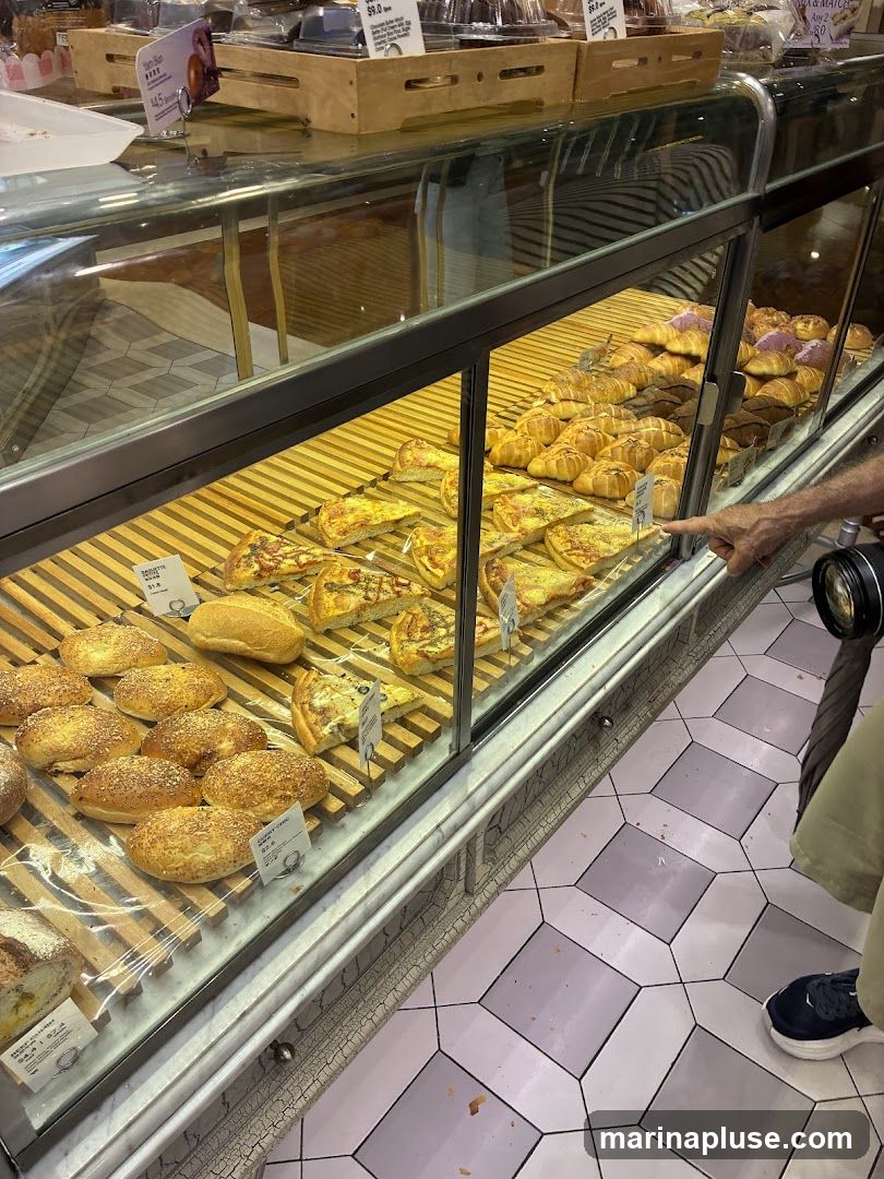 A display case filled with various baked goods, including croissants, pizzas, and bread rolls. A person's hand is visible pointing at one of the items. The floor has a geometric tile pattern.