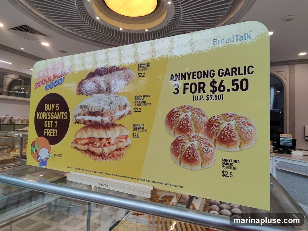 A yellow promotional sign from BreadTalk displays various korissant flavors and a special offer for Annyeong Garlic bread. The sign features images of the food items and their prices. It also includes a "Buy 5 Korissants, Get 1 Free" offer and a "3 for $6.50" deal on Annyeong Garlic bread. The promotion is valid until August 15, 2021.