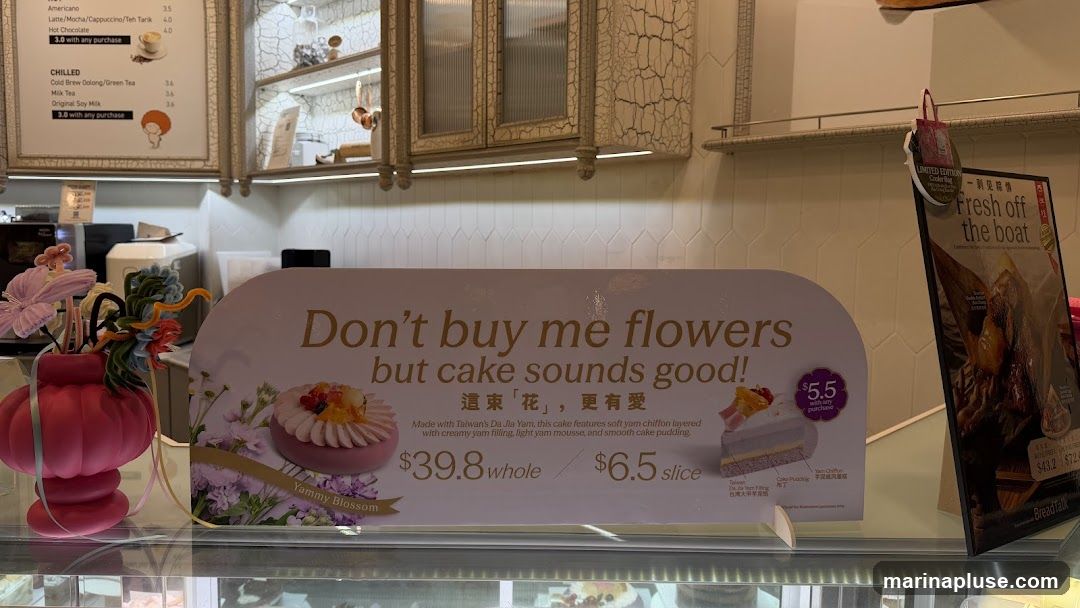 A sign advertising a cake with the slogan "Don't buy me flowers but cake sounds good!". The cake is described as being made with Taiwan's Da Jia Yam and features soft yam chiffon layered with creamy yam filling, light yam mousse, and smooth cake pudding. The price for a whole cake is $39.8 and a slice is $6.5. There is also a smaller promotion for $5.5 with any purchase. A menu board with coffee and tea options is visible in the background.