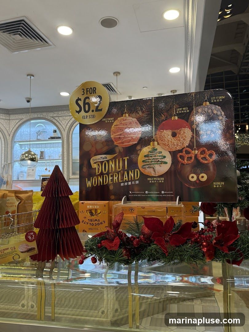 A display featuring donuts with a holiday theme, branded as 'Donut Wonderland' by BreadTalk. A promotional sign advertises '3 for $6.2' (U.P. $7.8). Several donuts are depicted, including 'Jingle Berry', 'Saint Nick', 'Let It Snow', 'Holly Pistachio', and 'Rudolph'. A red paper Christmas tree and festive greenery with red flowers are also part of the display.