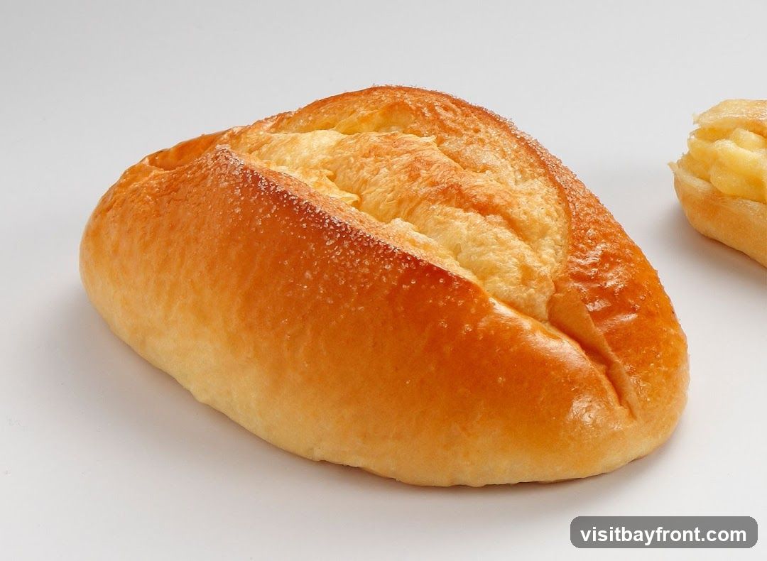 A close-up studio shot of a golden-brown bread roll with a slightly cracked top, dusted with sugar crystals. Another pastry, partially visible on the right, appears to be filled with a creamy yellow substance.