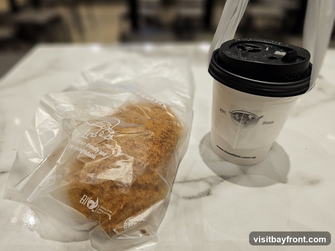 A white paper cup with a black lid, branded with the Toastbox logo and website, sits next to a plastic-wrapped pastry topped with shredded meat. The cup has "EST. 2005" and "www.toastbox.com.sg" printed on it. The plastic bag for the pastry has eco-friendly and biodegradable markings.