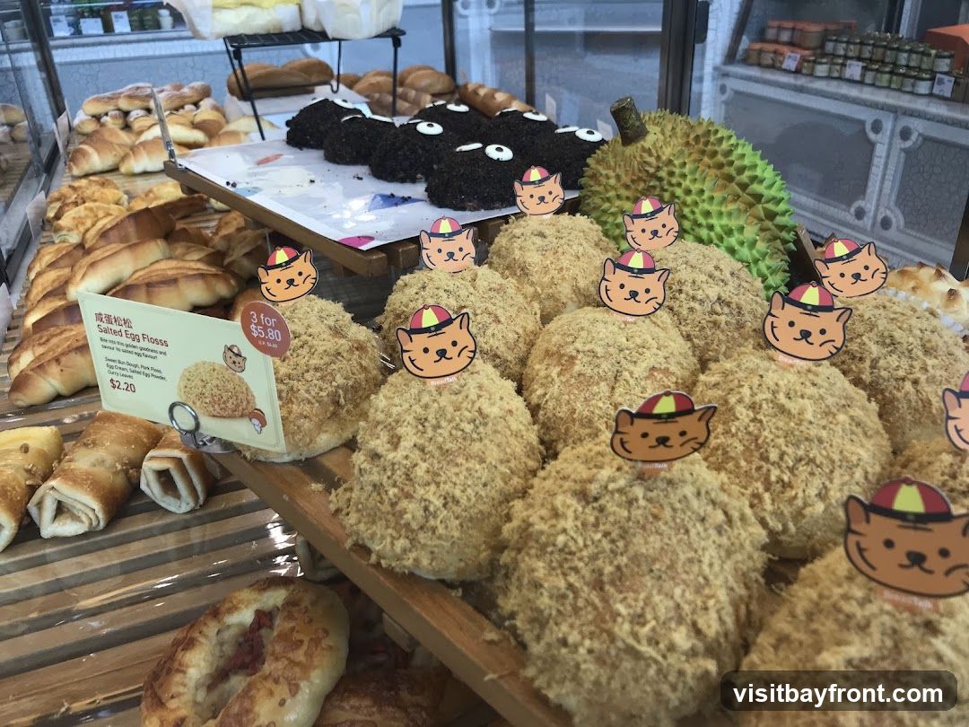 A close-up view of a bakery display case filled with various baked goods. In the foreground, several round buns covered in shredded meat or pork floss are visible, each adorned with a small cartoon cat sticker. To the left, a sign details "Salted Egg Floss" buns, priced at $2.20 each or 3 for $5.80. Behind these, rows of croissants and other pastries are arranged. In the background, dark, round pastries with white decorations and a durian fruit are also displayed. The overall impression is of a well-stocked and appealing bakery selection.