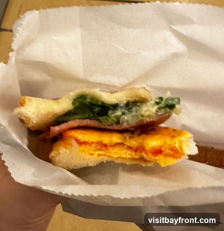 A close-up shot of a sandwich cut in half, revealing layers of ham, greens, and what appears to be scrambled eggs or cheese. The sandwich is wrapped in white paper.