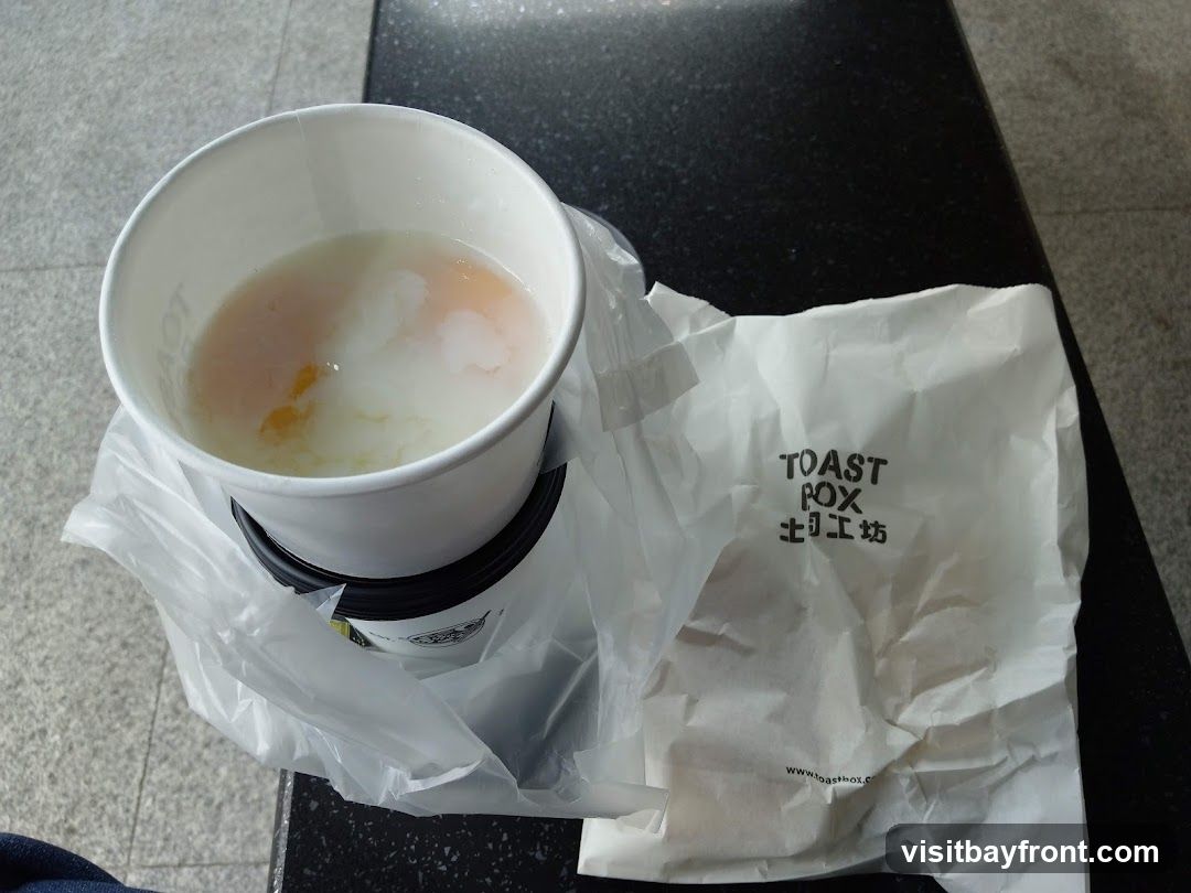 A white paper cup filled with soft-boiled eggs and broth sits inside a plastic bag. Next to it is a crumpled white paper bag with the logo "TOAST BOX" and Chinese characters printed on it. The website "www.toastbox.com" is visible at the bottom of the paper bag.