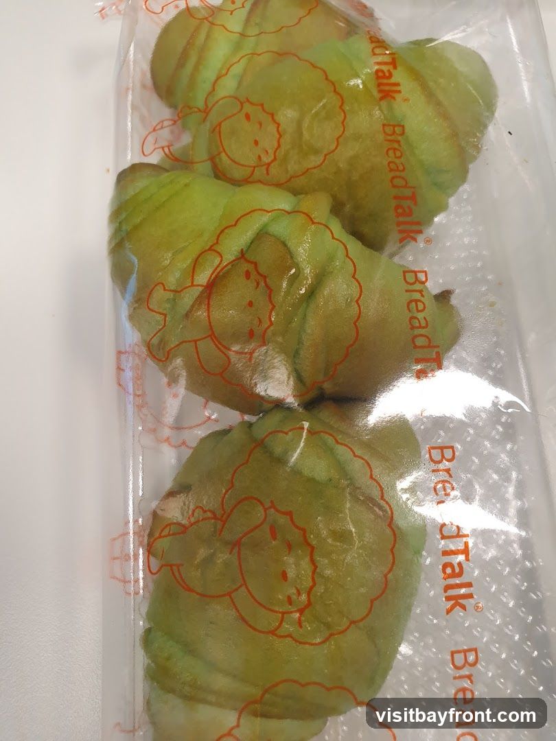 Three green croissants are packaged in a clear plastic bag. The bag has an orange outline of a sheep printed on it, and the words "BreadTalk" are printed vertically in orange.