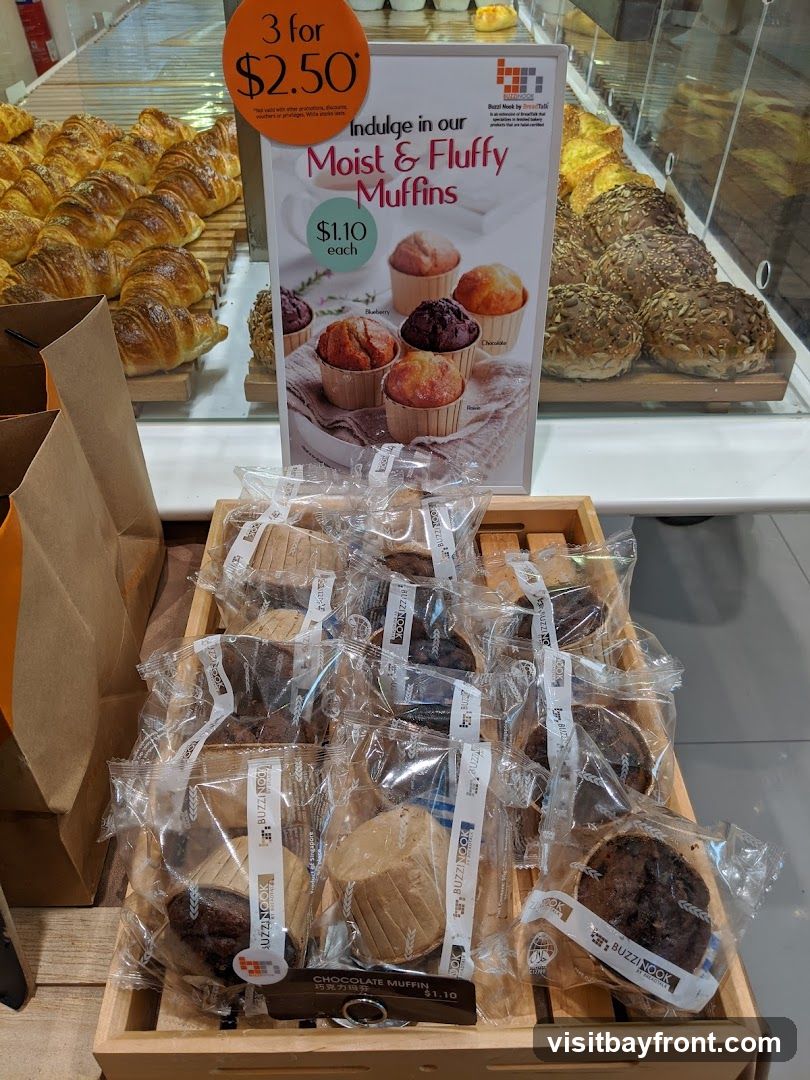 A display of baked goods including rows of croissants and various muffins. A sign advertises "Moist & Fluffy Muffins" with a promotion of "3 for $2.50" or "$1.10 each". Individual muffins are packaged in plastic.