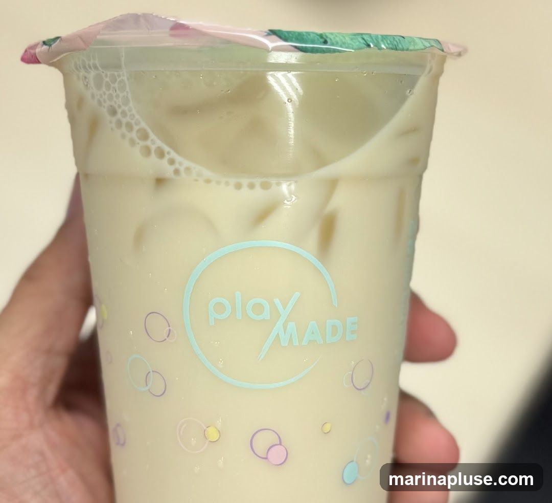 A close-up shot of a light-colored beverage in a clear plastic cup, held by a hand. The cup has a colorful, patterned lid and features a circular logo with the text "play MADE" in light blue. The cup itself is decorated with small, colorful circles and bubbles. The drink appears to contain ice and has a frothy layer at the top.