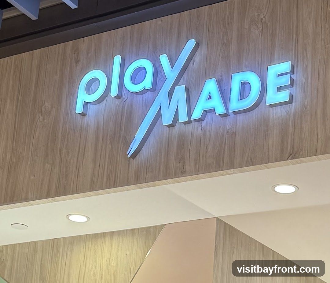 A close-up shot of a glowing blue sign that reads "play MADE" against a wood-paneled background. The sign is illuminated from behind, creating a soft glow. The letters are three-dimensional and appear to be made of acrylic or a similar material. The background has a light wood grain texture. Below the sign, there is a white soffit with recessed lighting.