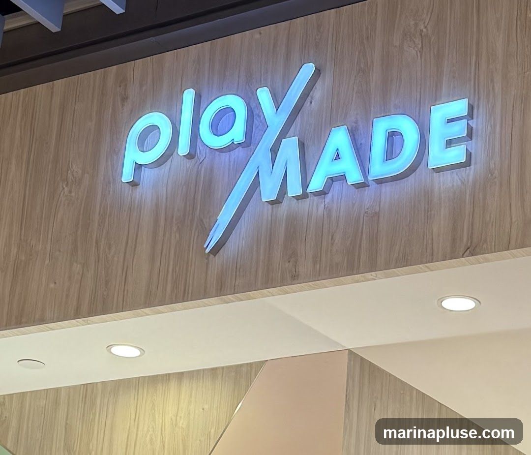 A close-up shot of a glowing blue sign that reads "play MADE" against a wood-paneled background. The sign is illuminated from behind, creating a soft glow. The letters are three-dimensional and appear to be made of acrylic or a similar material. The background has a light wood grain texture. Below the sign, there is a white soffit with recessed lighting.