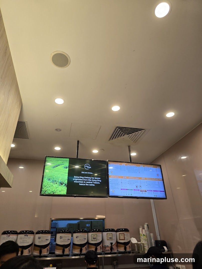 A view of a tea shop counter with two large digital screens displaying information and menus. Below the screens are several large beverage dispensers. The left screen shows a scenic image of tea fields and text about Dong Ding Oolong tea. The right screen displays a menu with various drink categories and prices. The dispensers are labeled with different drink options and sizes.