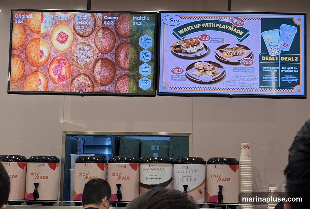Two digital menu boards are displayed above a row of drink dispensers. The left board shows various flavors of what appear to be small pastries or balls, including Original ($3.4), Pink Cactus Boba ($4.5), Black Sesame ($4.2), Cocoa ($4.2), and Matcha ($4.2). The right board advertises breakfast items and deals, with options like Scallion Egg Egget ($4.5), Ham & Cheese Pancake ($5.2), and Chicken Luncheon Meat Scallion Pancake ($5.2). It also promotes two deals for topping up M-Milk Tea or M-Classic Tea. Below the menus, several drink dispensers branded with "play MADE" are visible.
