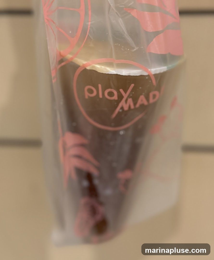 A close-up shot of a drink in a clear plastic bag. The bag has pink tropical leaf and citrus fruit designs. The logo "play MAD" is visible on the cup inside the bag.