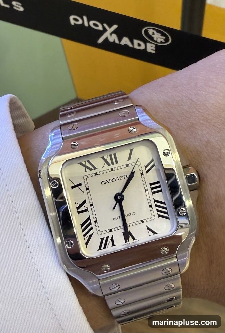 A close-up shot of a Cartier Santos watch on a person's wrist. The watch has a silver metal band and a white face with Roman numerals. The word "CARTIER" is visible on the face, along with "AUTOMATIC" and "SWISS MADE". In the background, a black band with the words "playXMADE" and a logo is visible, along with colorful geometric shapes.