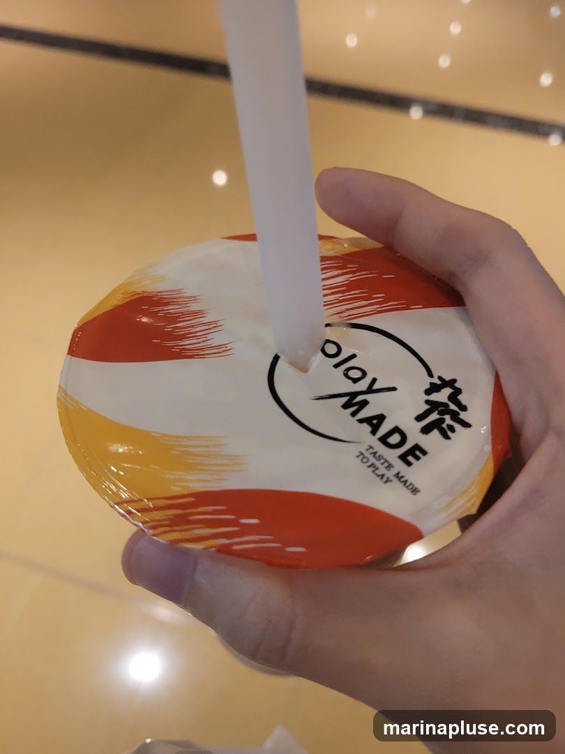 A hand holds a drink cup with a sealed lid. The lid has a colorful design with red, yellow, and white brush strokes. A black circular logo with text reads "play MADE" and some Chinese characters. Below that, it says "TASTE MADE TO PLAY". A white straw is inserted through the lid.