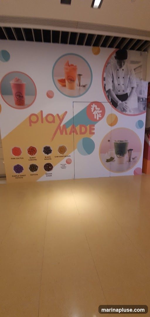A large display board with the text "play MADE" and images of drinks and toppings. The board features two circular images of colorful beverages, one pink and one orange, with garnishes. Another circular image shows a green beverage with ice. A black and white image of a chef preparing food is also visible. Below the text, there are circular images of various toppings with their names: Pink Cactus, Burnt Caramel, Black Sesame, Chrysanthemum, Purple Sweet Potato, Matcha, and Brown Sugar. A smaller text box indicates "Add Tiny Tapioca Pearls". The display is set against a white background with colorful circles.