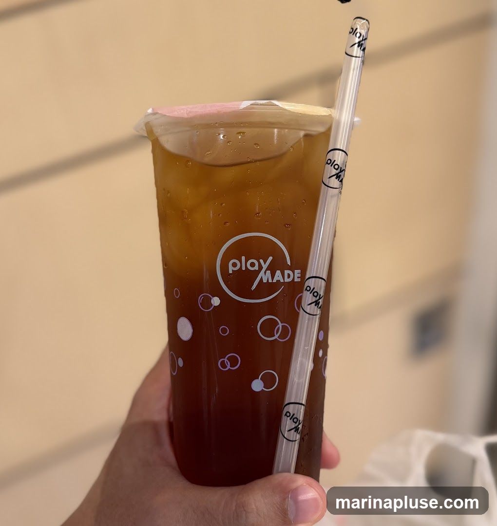 A hand holds a clear plastic cup filled with iced tea. The cup has a pink-colored seal and is decorated with a repeating pattern of circles and the Playmade logo. A clear plastic straw with the Playmade logo printed on it is inserted into the drink. The background is a light-colored, textured wall.