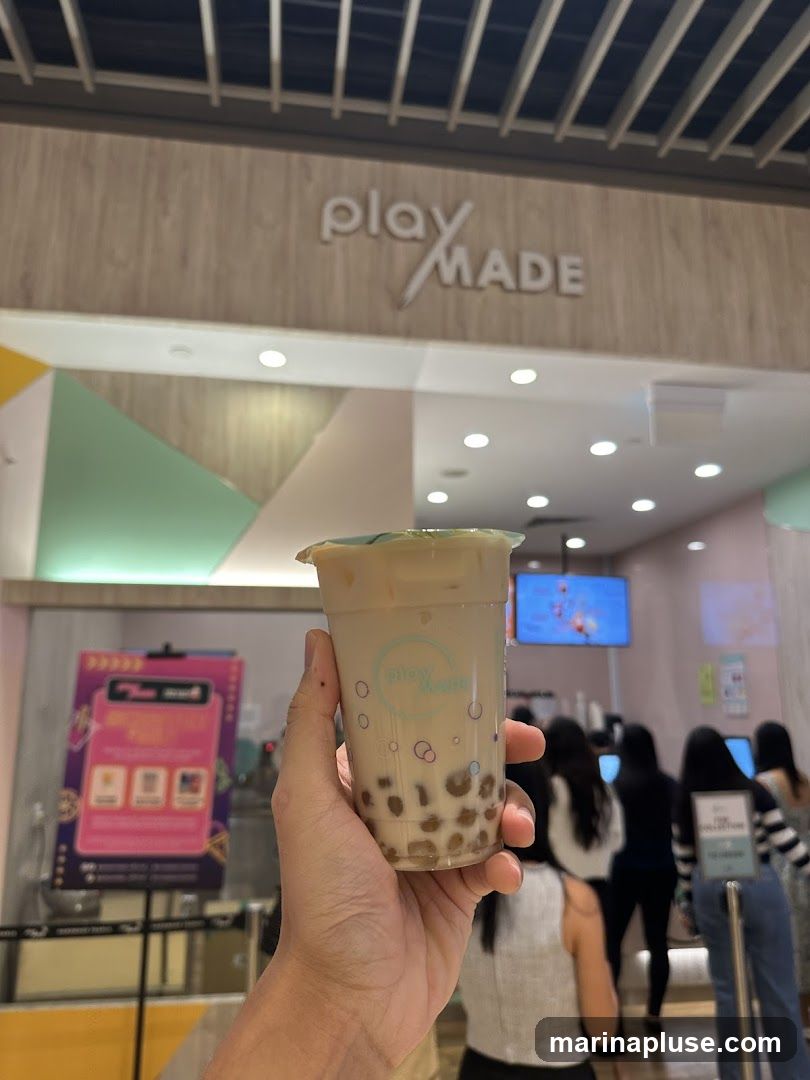 A hand holds a plastic cup of bubble tea in front of a store with a sign that says "play MADE". The drink has a light brown color and contains tapioca pearls at the bottom. The store interior is visible in the background, with people queuing and a screen displaying content.