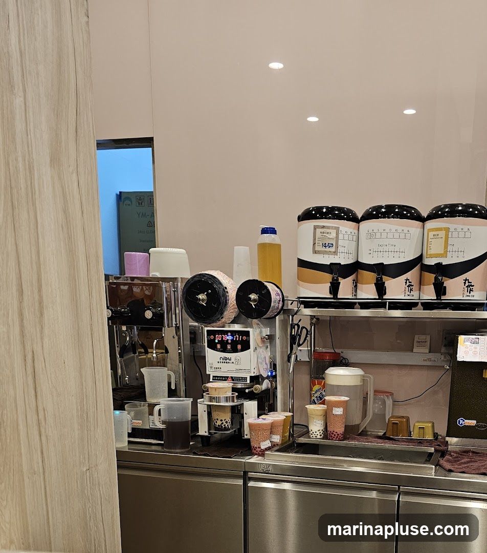 A beverage preparation station with multiple dispensers, a sealing machine, and various cups and ingredients. Three large dispensers with black lids and tan and black accents are visible, each with a tap. A sealing machine with a digital display is in the center, with cups of prepared beverages in front of it. Several plastic pitchers and bottles of liquid are also present. The overall scene suggests a bubble tea or similar beverage shop.