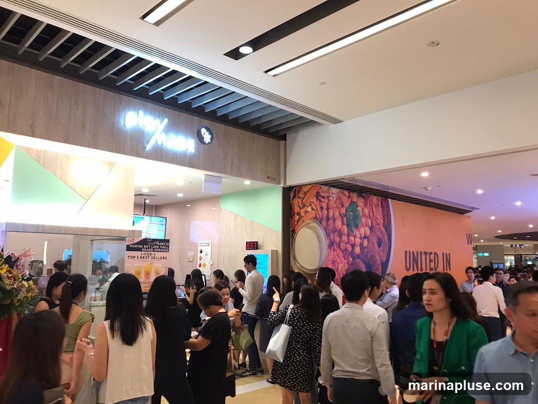 A crowd of people are gathered in front of a food outlet with a prominent sign that reads "playmade". There is a large promotional banner on the right side of the image featuring a colorful array of food items and the text "UNITED IN". A digital display shows the time as 8:37. Several customers are queuing, suggesting a popular establishment.