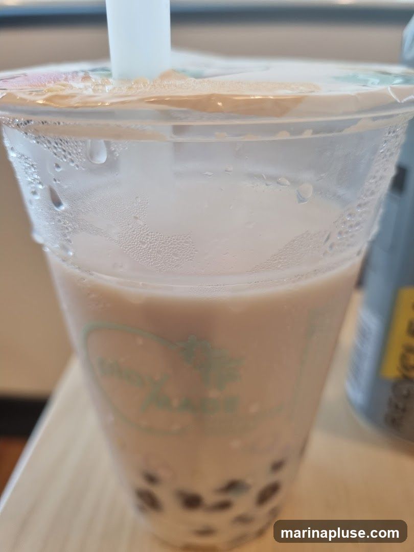 A close-up shot of a bubble tea drink in a clear plastic cup. The cup has condensation on the outside and a white lid with a straw. The drink appears to be a milky brown color with tapioca pearls at the bottom. A logo is visible on the cup, but it is partially obscured and difficult to read. A gray can with a yellow label is visible in the background.