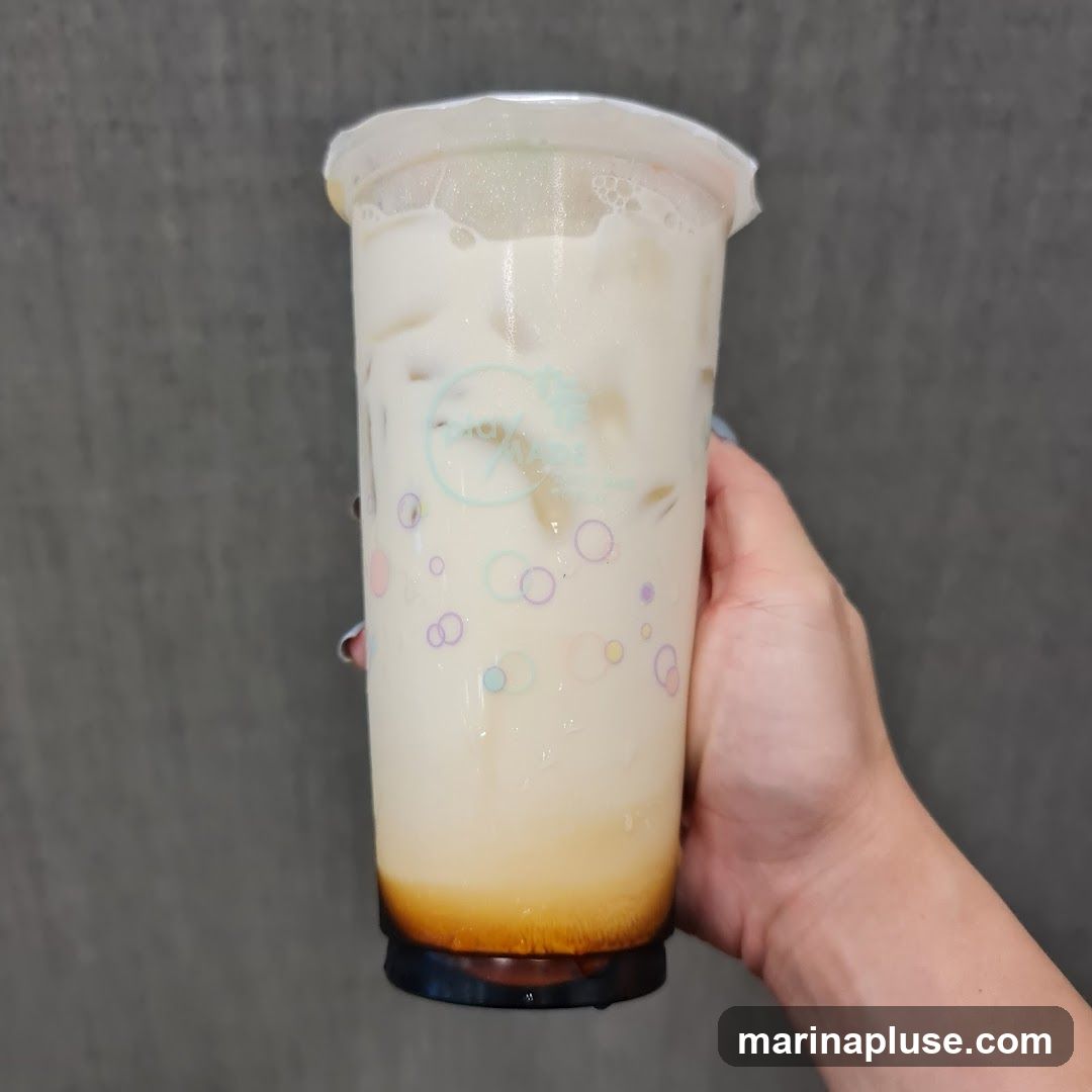 A hand holds a clear plastic cup filled with brown sugar milk tea. The drink has a layer of brown sugar syrup at the bottom, topped with milk and ice. The cup has a logo and colorful bubble designs.