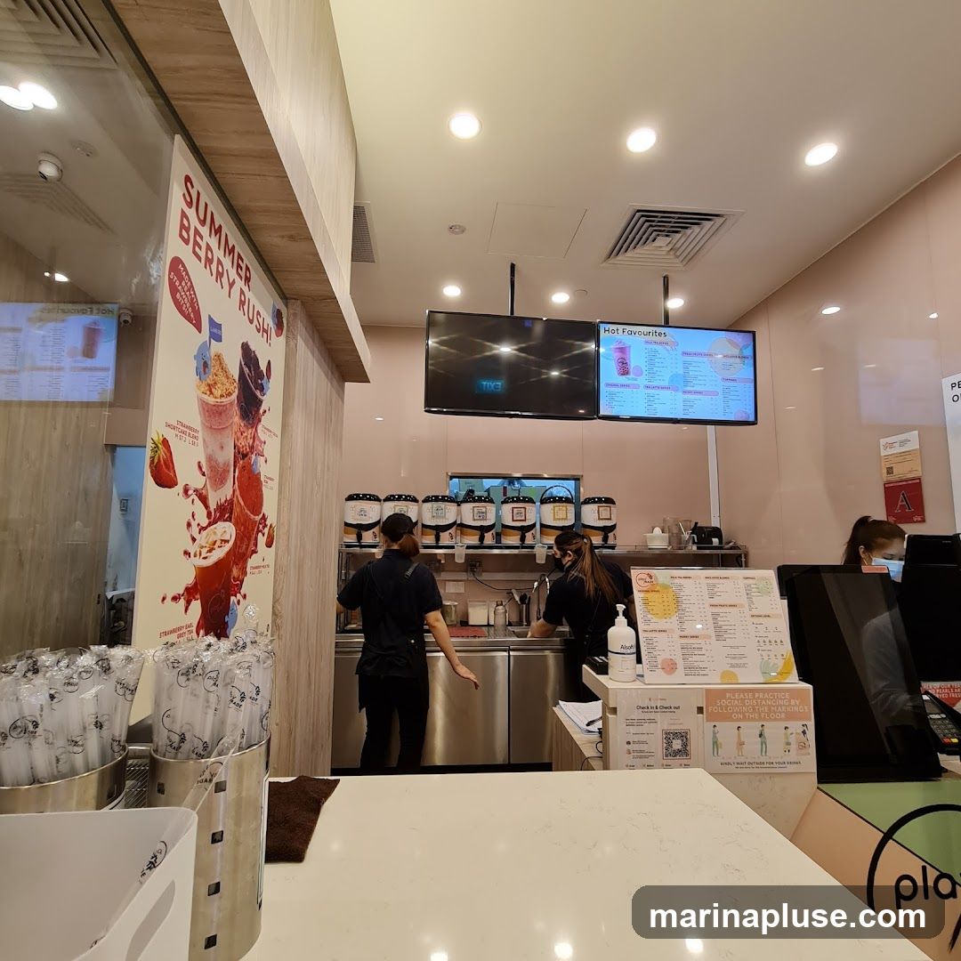 A view of the counter area in a bubble tea shop. Two employees are visible behind the counter, preparing drinks. A large promotional poster for a "Summer Berry Rush" is on the left. Digital menu boards are mounted on the wall above the counter, displaying "Hot Favourites" and other drink options. Several dispensers for toppings or ingredients are lined up behind the counter. In the foreground, there are containers of straws and a white countertop.