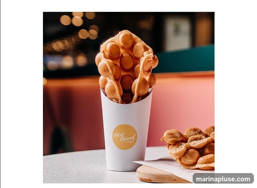 A close-up shot of a bubble waffle in a white paper cup with a yellow circular logo that says "play ground by mygout". Another bubble waffle is visible on the right side of the image. The background is blurred with bokeh lights and pink and teal colors.