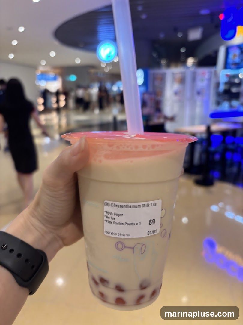 A hand holds a cup of Chrysanthemum Milk Tea with pink cactus pearls. The cup has a label indicating "25% Sugar", "No Ice", and "Pink Cactus Pearls x 1". The background shows a blurred indoor shopping mall environment with people and bright lights.