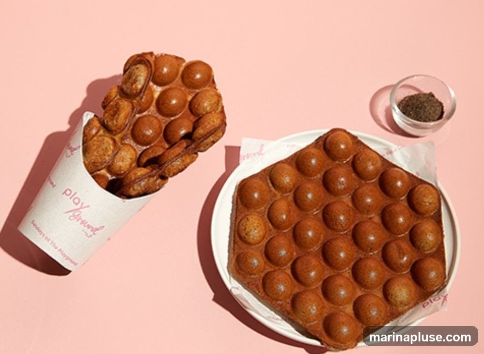 Two bubble waffles are displayed on a pink background. One waffle is served in a paper cone with the logo "play / Ground" visible. The other waffle is on a white plate. A small glass bowl of dark powder is to the right of the plate.