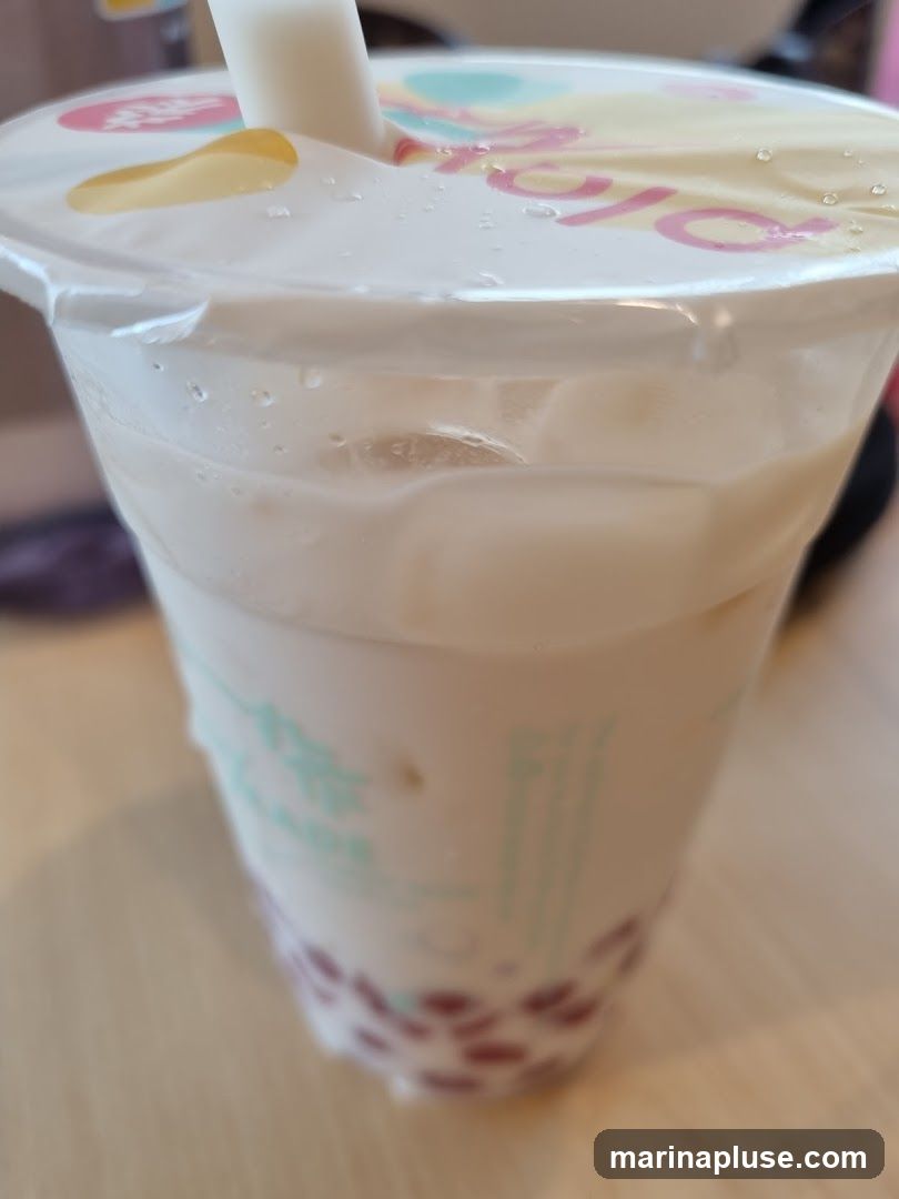 A close-up shot of a bubble tea drink in a clear plastic cup. The cup has a white lid with a pink logo and a white straw. The drink itself is a creamy white color with ice cubes and tapioca pearls at the bottom. The cup has some green text and a pattern of red dots near the base. The drink is on a wooden surface.