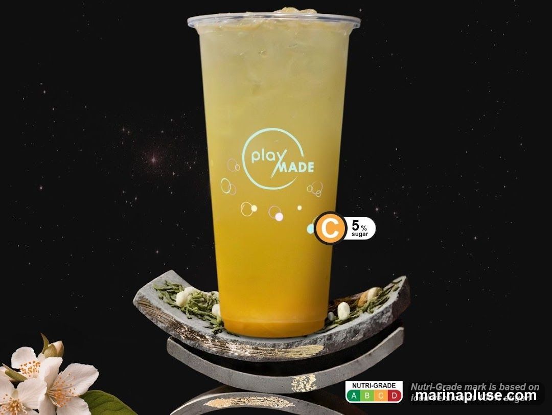 A tall plastic cup filled with a yellow-orange beverage, likely a tea or juice, with ice. The cup has the "play MADE" logo and a "C 5% sugar" nutritional grade. The drink is presented on a decorative stand with what appears to be tea leaves and nuts. A sprig of jasmine flowers is visible in the lower left corner. The background is dark with scattered white specks resembling stars.