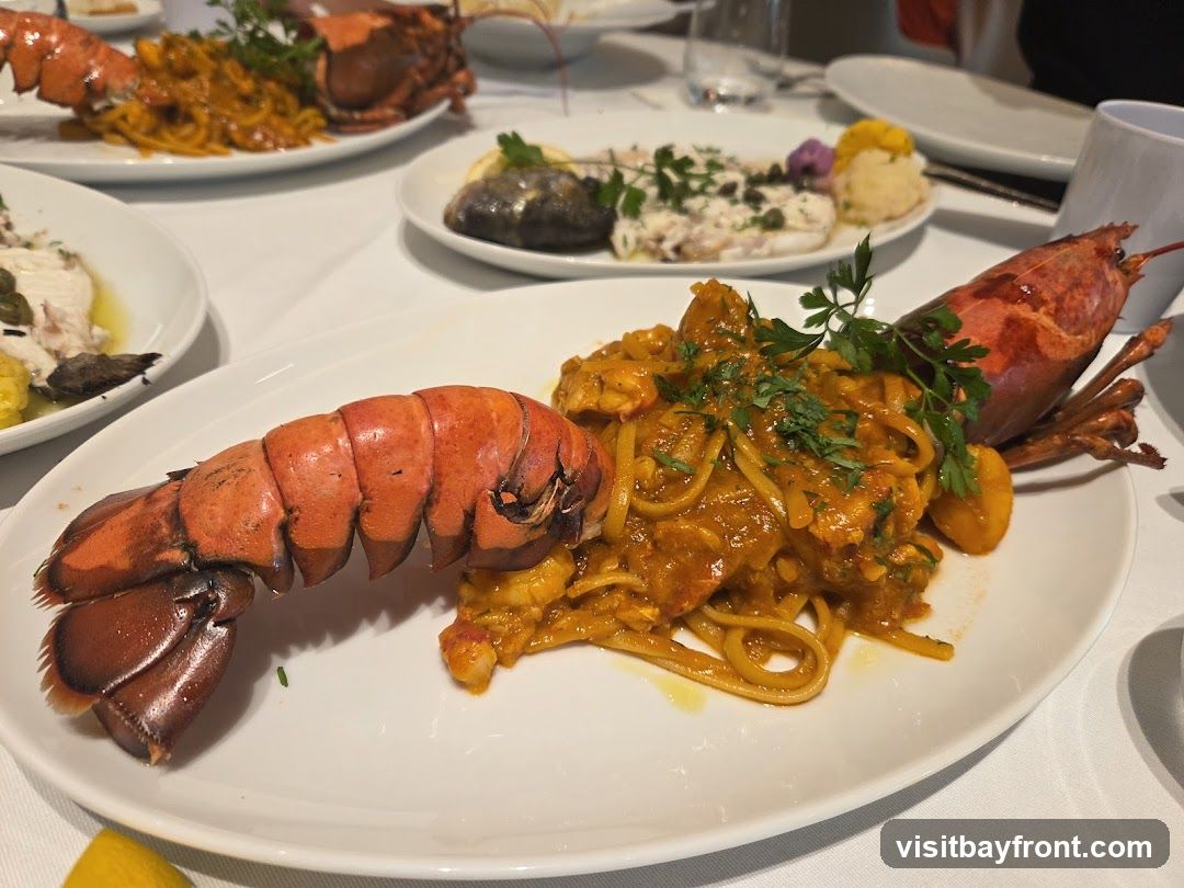 A close-up shot of a white plate holding a lobster tail served with pasta in a rich orange sauce. The pasta appears to be linguine, and it is garnished with fresh green herbs. In the background, other plates of food are visible, including what looks like a fish dish and another lobster entree. The setting appears to be a restaurant with white tablecloths.