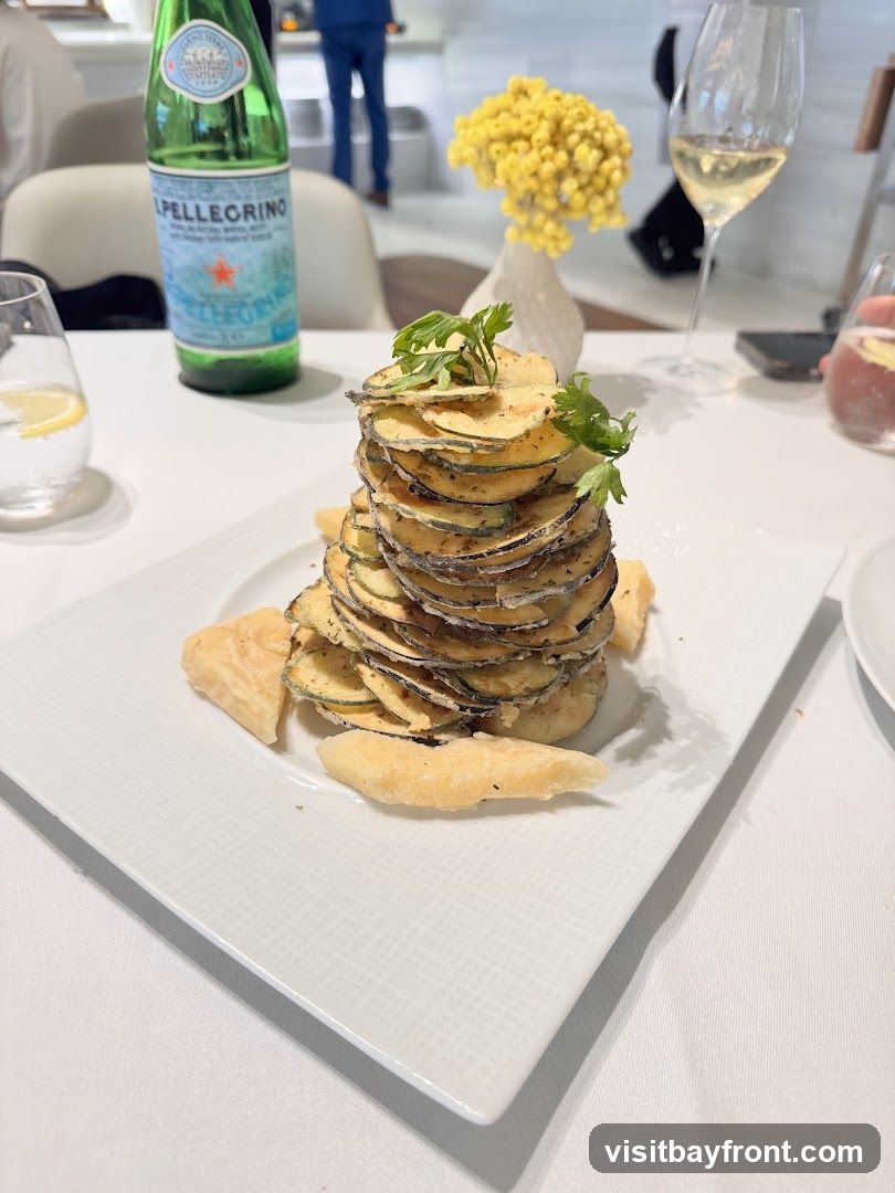 A tall stack of thinly sliced, fried zucchini chips is presented on a white rectangular plate. The zucchini is seasoned with herbs and spices. Two pieces of fried dough or cheese are placed on the side of the stack. In the background, a bottle of San Pellegrino water, a glass of white wine, and a small vase with yellow flowers are visible on a white tablecloth. The setting appears to be a restaurant.