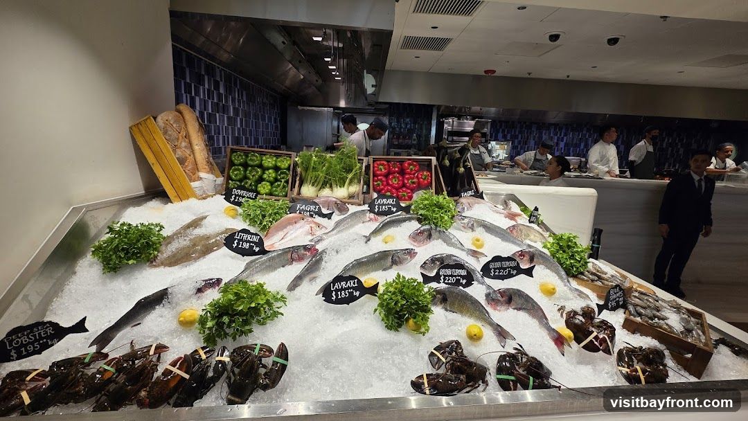 A display of fresh seafood on ice, including lobsters, various fish, and some vegetables. Chefs and staff are visible in the background in a kitchen setting.
