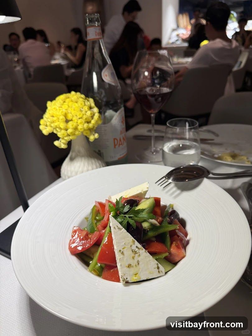 A close-up view of a Greek salad served in a white bowl on a table at a restaurant. The salad contains tomatoes, cucumbers, green peppers, olives, and feta cheese, garnished with parsley and drizzled with olive oil. A bottle of San Pellegrino water, a glass of red wine, and a glass of water are also visible on the table. In the background, other diners are seated at tables, and the restaurant's interior is dimly lit.
