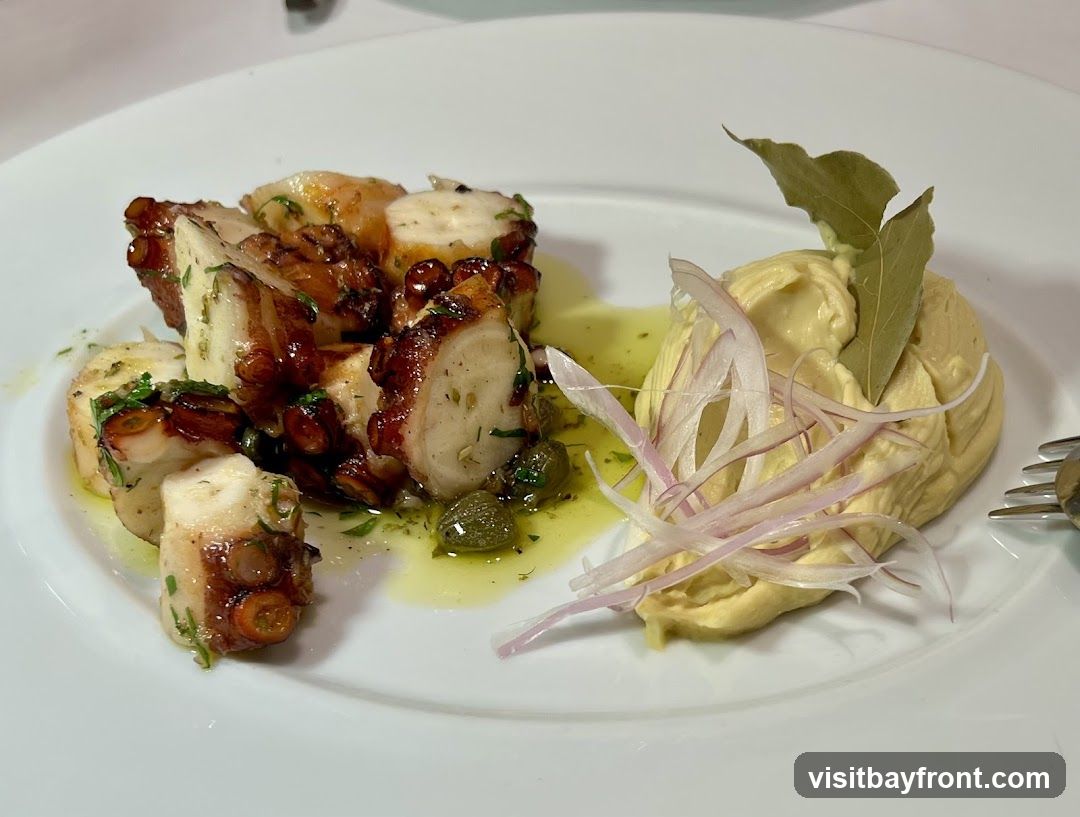 A white plate holds a serving of grilled octopus tentacles, seasoned with herbs and capers, and drizzled with olive oil. To the side is a dollop of hummus, garnished with thinly sliced red onion and bay leaves. A fork is visible on the right side of the plate.