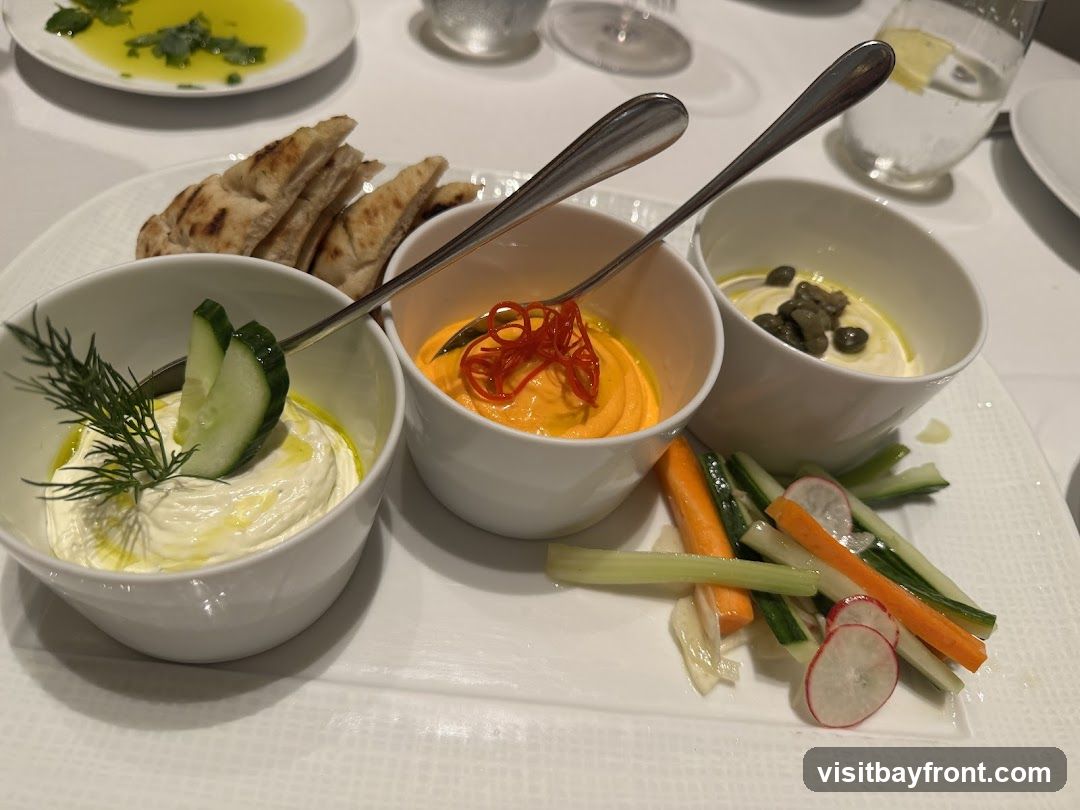A platter of three dips served with grilled pita bread and crudités. The dips appear to be a white creamy dip with dill and cucumber, an orange dip topped with red pepper strips, and a lighter colored dip topped with capers. The crudités include carrot sticks, cucumber sticks, celery sticks, and radish slices. A small dish of olive oil with herbs is visible in the background.