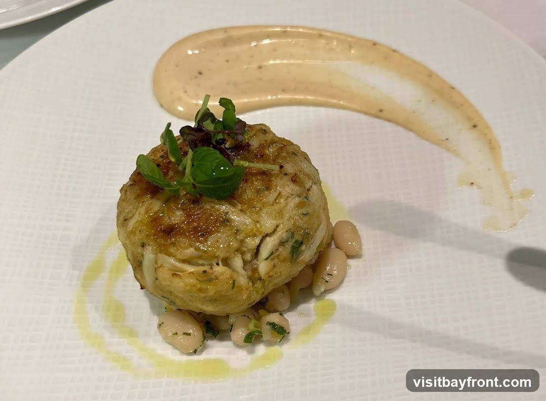 A close-up shot of a single crab cake served on a white plate. The crab cake is garnished with microgreens and sits atop a bed of white beans. A swirl of creamy sauce is artfully placed on the side of the plate.