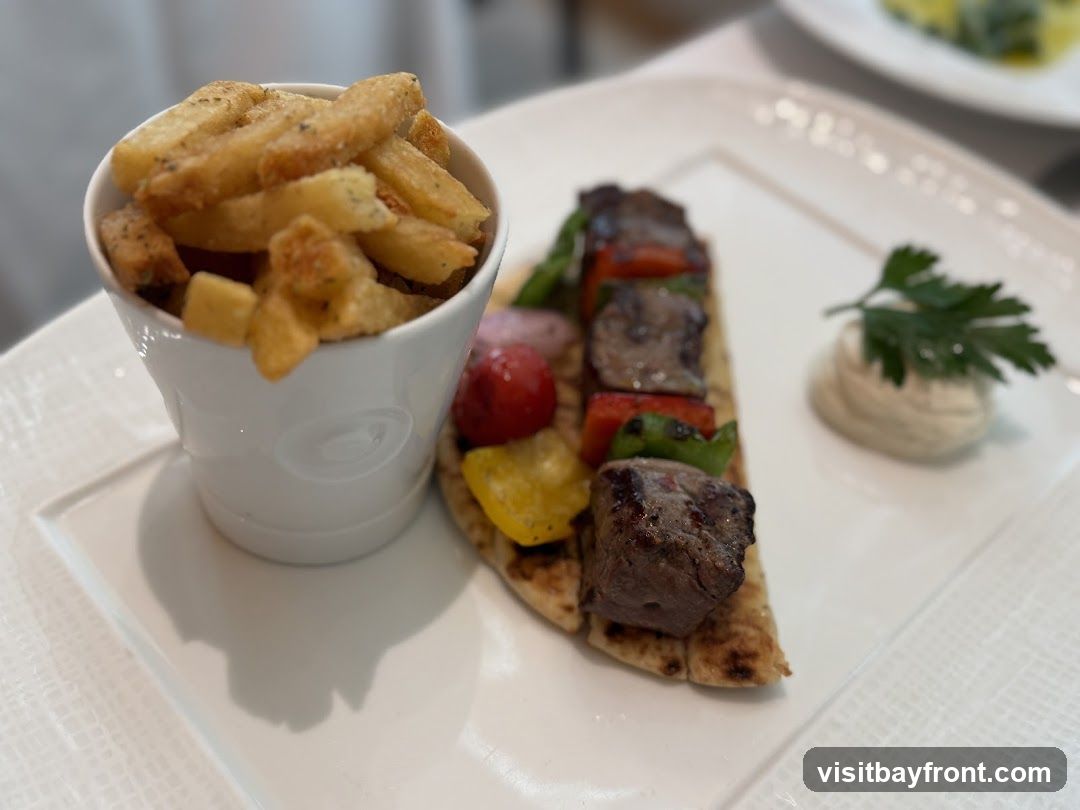 A plate of food featuring beef skewers with bell peppers and tomatoes served on pita bread, accompanied by a side of seasoned fries in a white ceramic cup and a dollop of dip garnished with parsley.