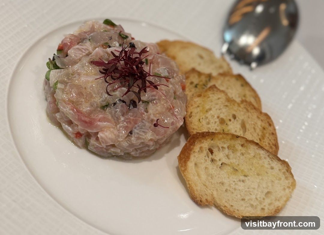 A close-up shot of a white plate featuring a mound of fish tartare, garnished with microgreens. The tartare appears to be mixed with finely chopped herbs and possibly capers or olives. Alongside the tartare are several slices of toasted baguette, lightly brushed with olive oil. In the blurred background, a shiny, dark object, possibly a spoon or a decorative element, is visible.