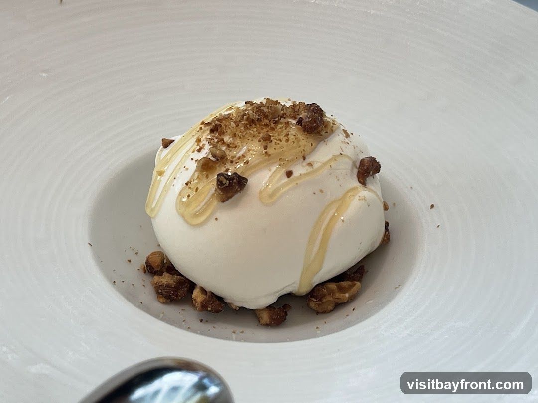A close-up shot of a white dessert, possibly ice cream or mousse, served in a white bowl with concentric ridges. The dessert is topped with chopped nuts and drizzled with honey. A spoon is partially visible in the foreground.